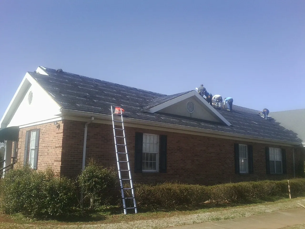 Crew tearing shingles off a brick ranch roof with a ladder set in Hubbardston